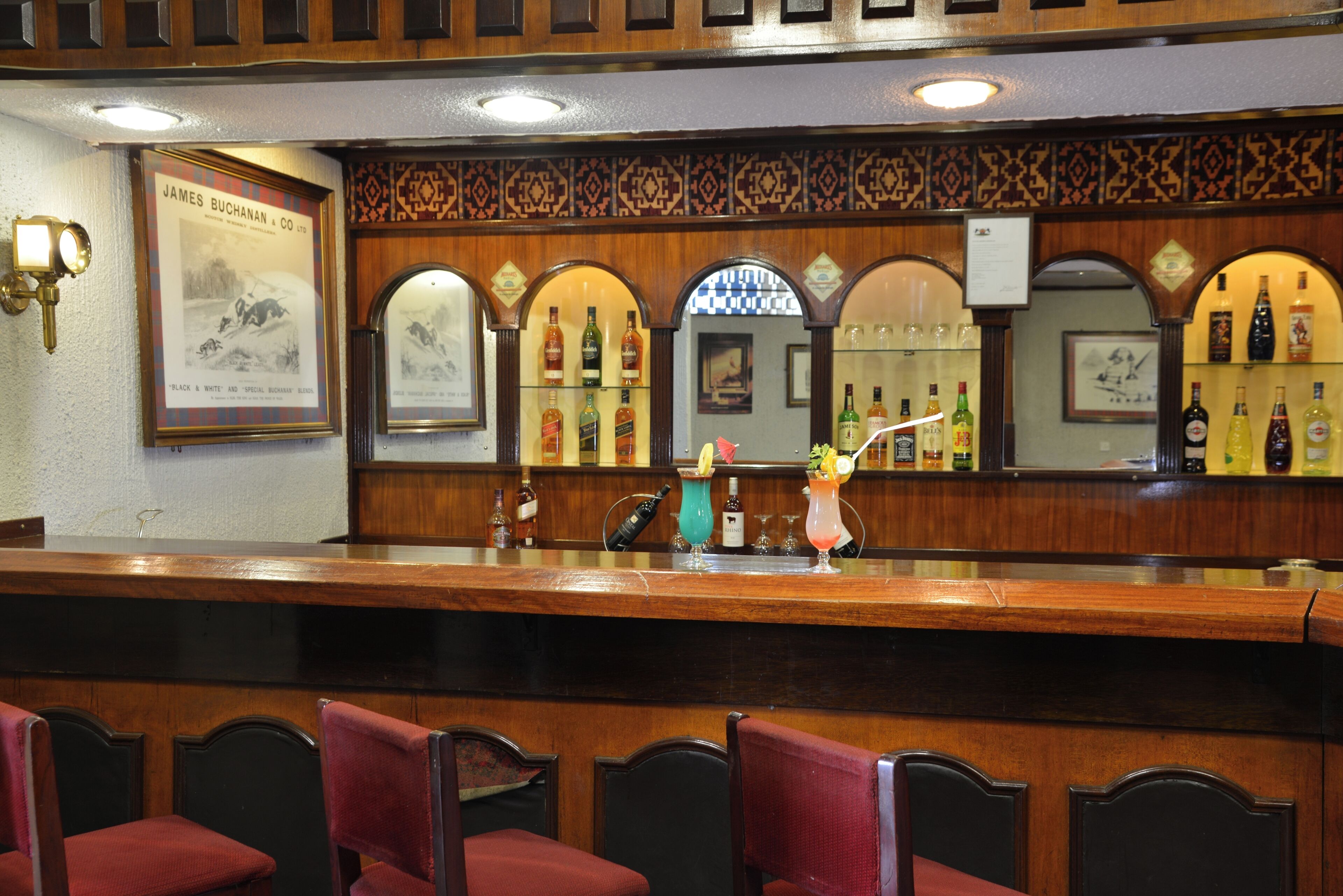 Zona de bar amb taulell de fusta, cadires tapissades de vermell i diverses ampolles de licors.