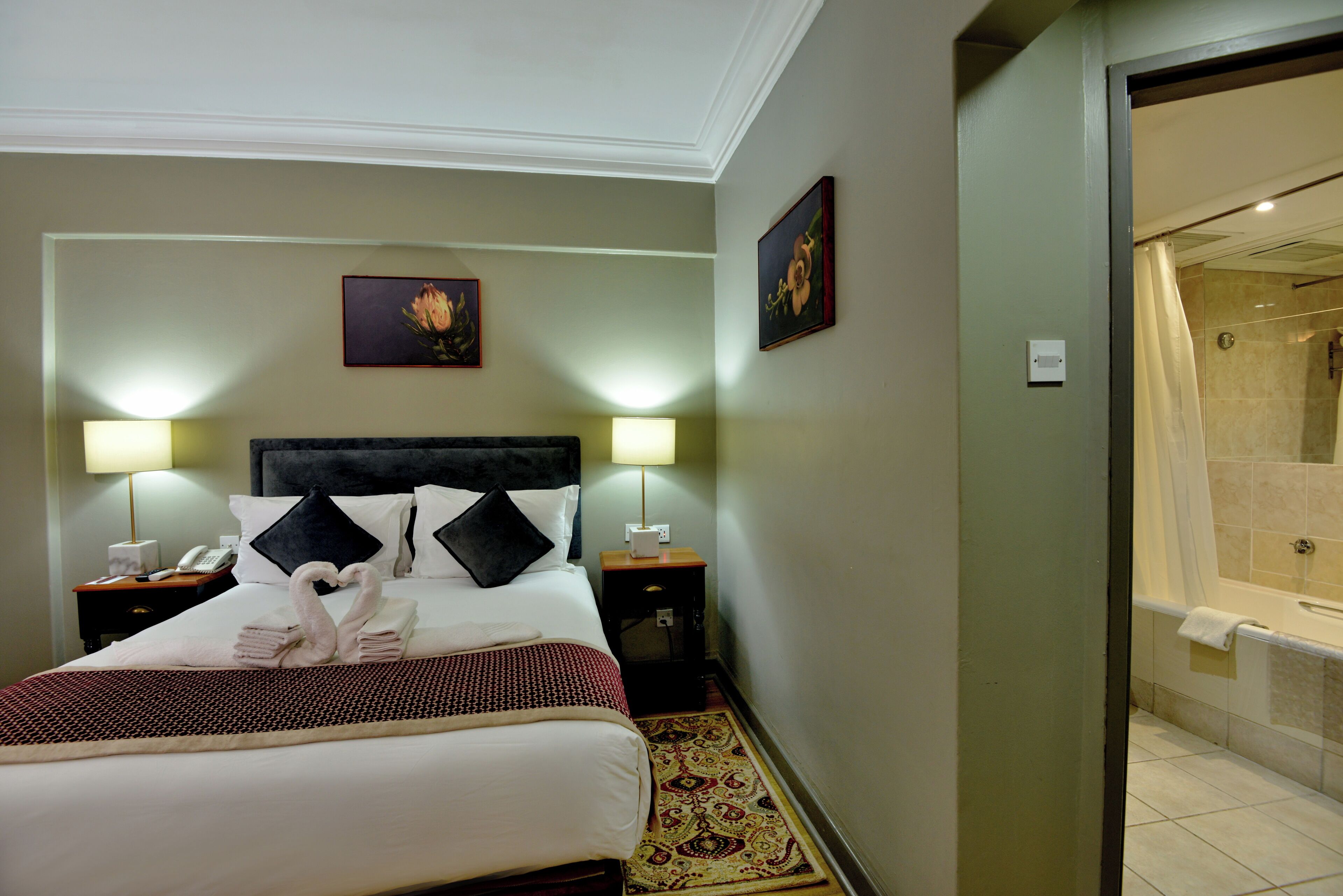 A hotel room with a bed, two bedside tables with lamps, a nightstand, a shower area, and a patterned rug.