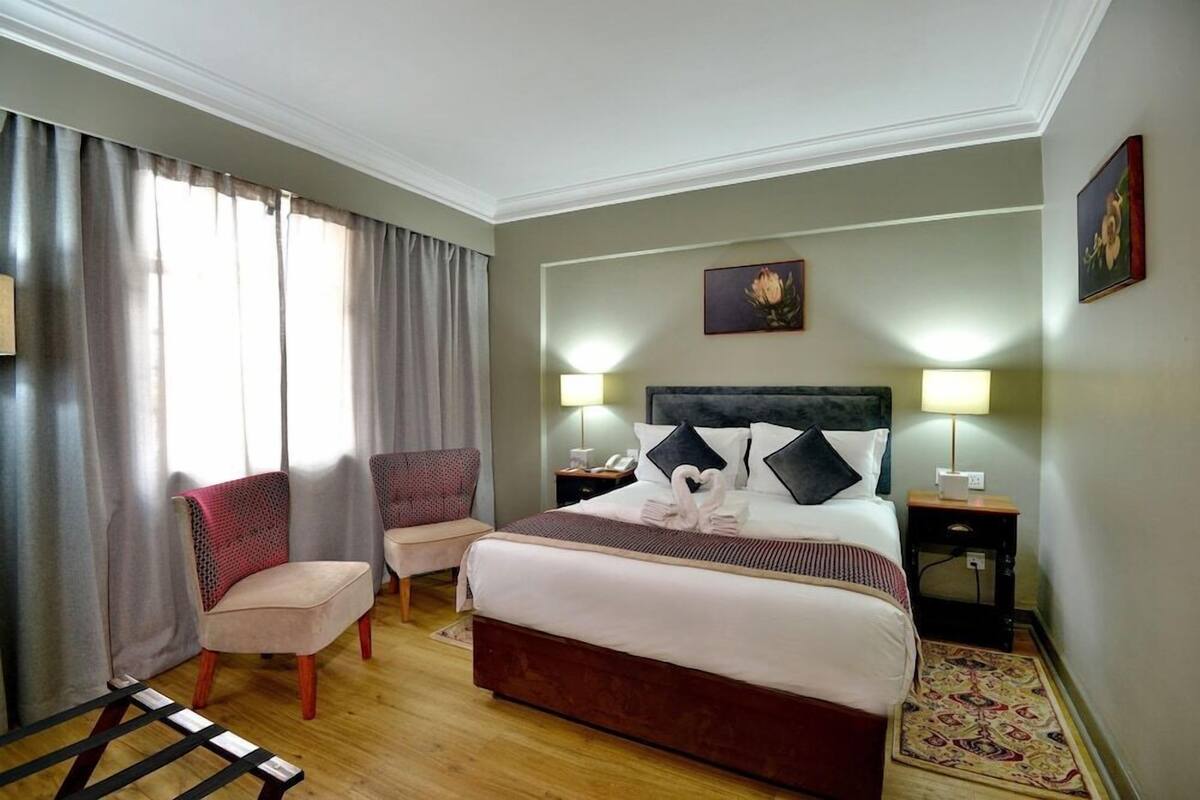 A hotel room with a bed, two chairs, a nightstand with a lamp, a wall-mounted picture, and a window with curtains.