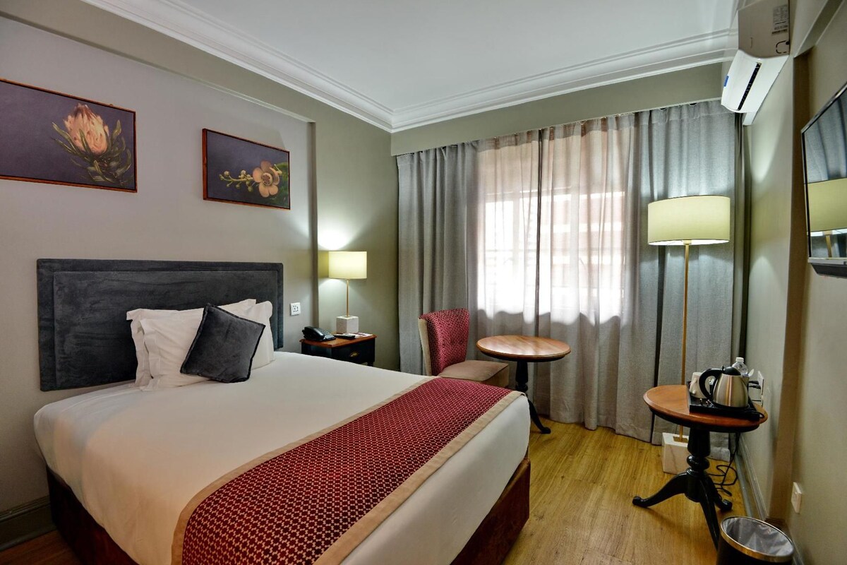 Une chambre d’hôtel avec un grand lit, une table de chevet avec une lampe, une chaise, une petite table ronde avec une bouilloire et un miroir au mur.
