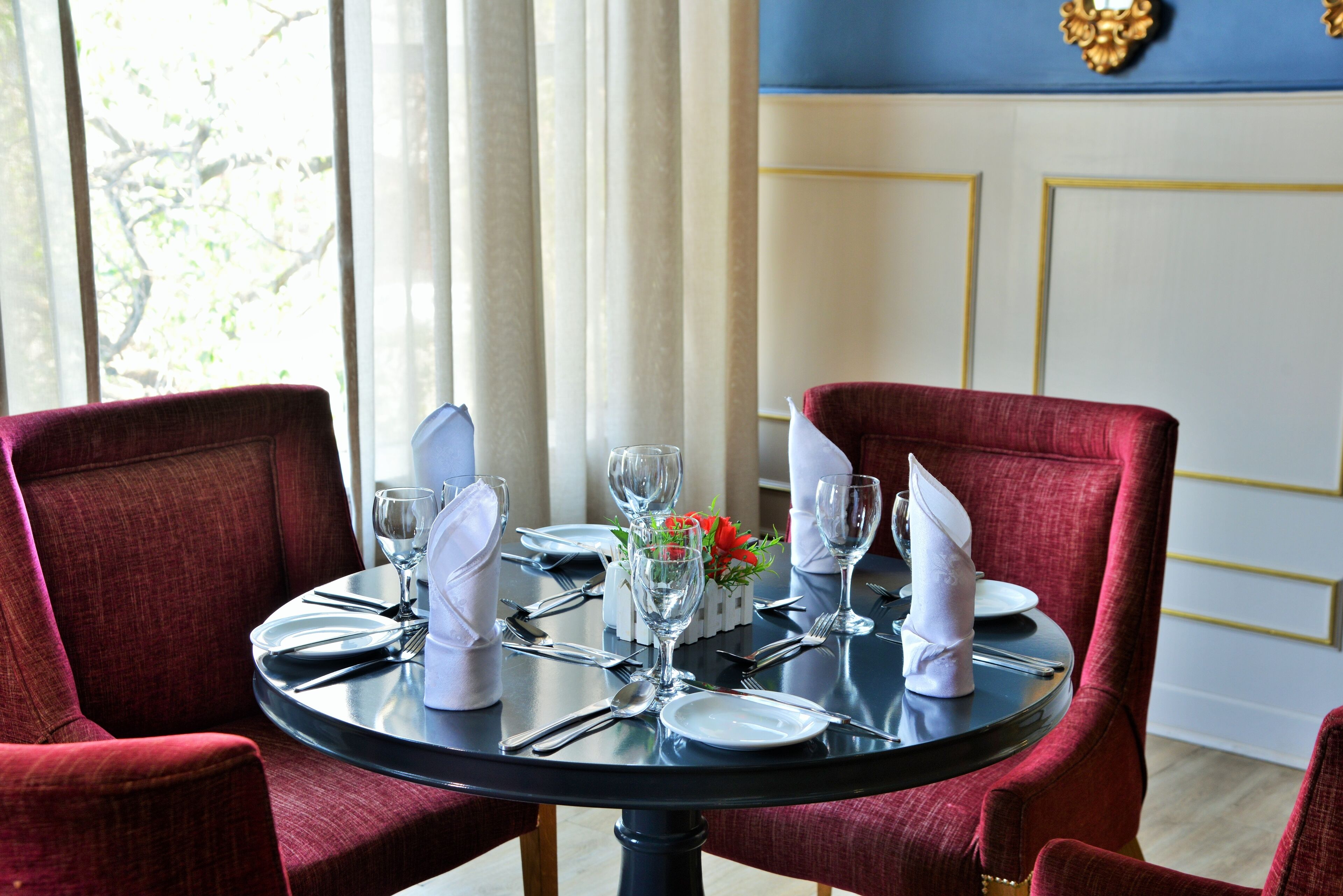 A dining table set for four with red upholstered chairs, white napkins, and a small vase with red flowers.