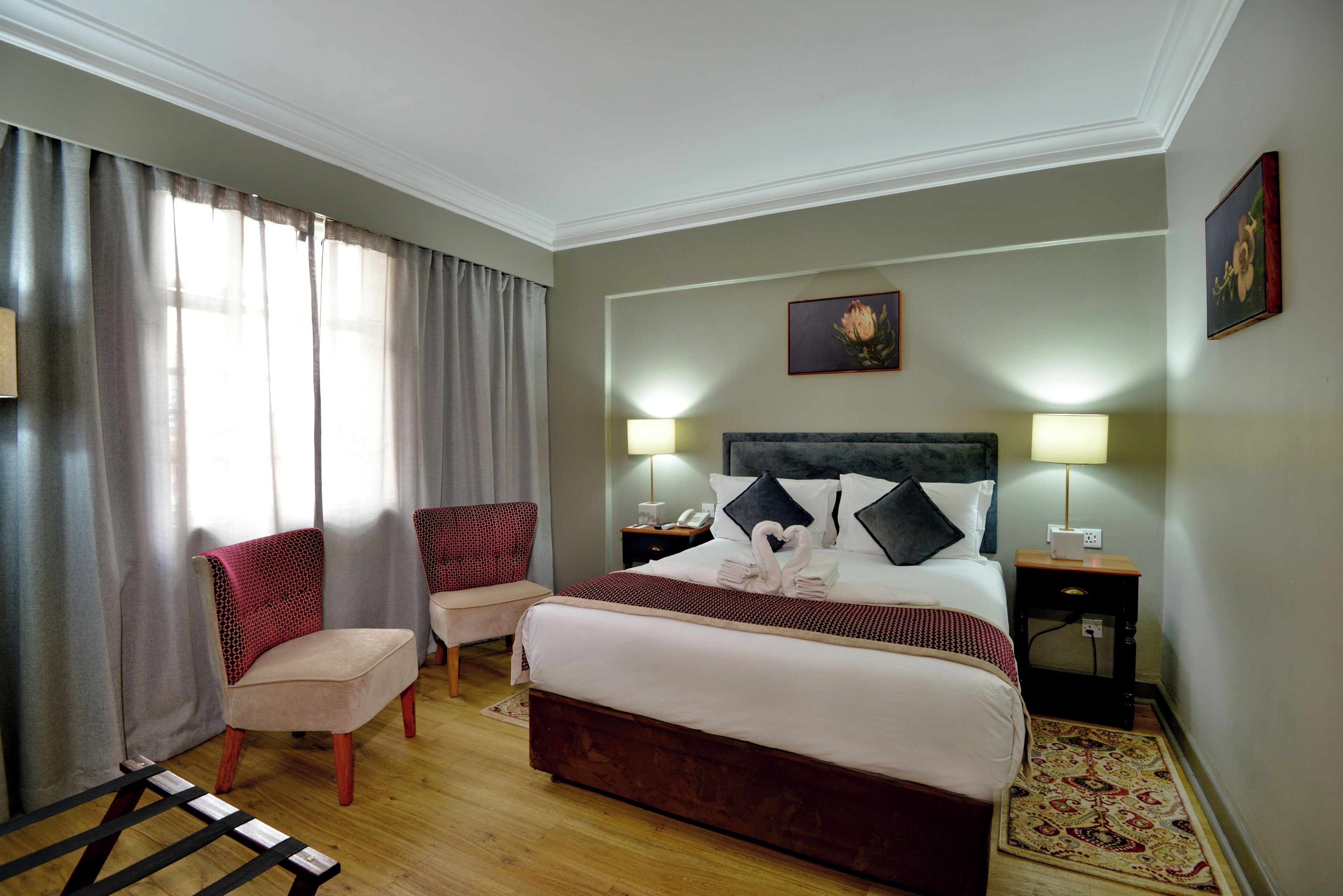 Una habitación de hotel con cama, dos sillas, una mesita de noche, una lámpara y un cuadro en la pared.