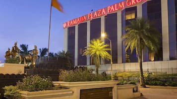 Ramada Plaza Palm Grove features a modern design with large windows and a flat roof, surrounded by palm trees and flowering plants.