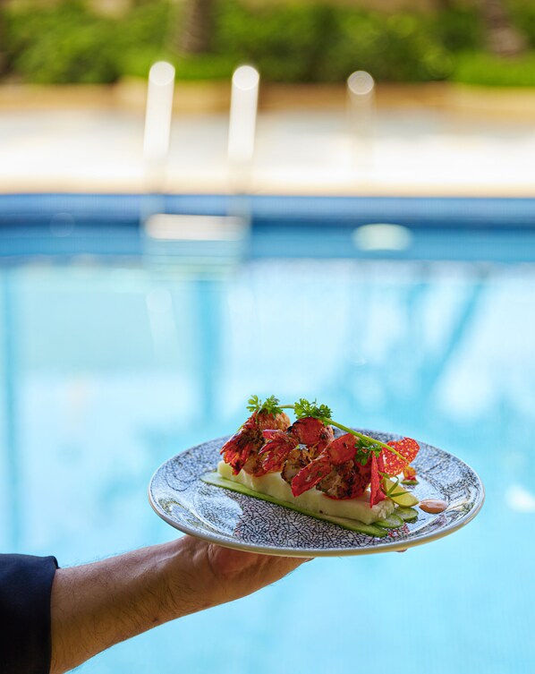 Una mano sostiene un plato con comida, una piscina y follaje al fondo.