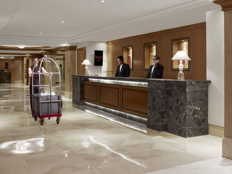 Uma área de recepção de hotel com balcão de mármore, carrinho de bagagem e funcionários na recepção.
