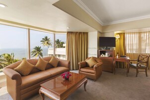 A hotel room with a large window offering a view of palm trees and the ocean. There is a sofa, a chair, a small table, and a TV.