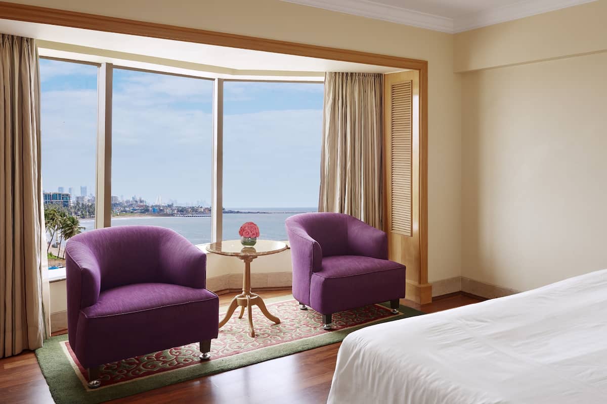 Une chambre d’hôtel avec une grande fenêtre, deux fauteuils violets, une petite table ronde avec un vase et un lit.