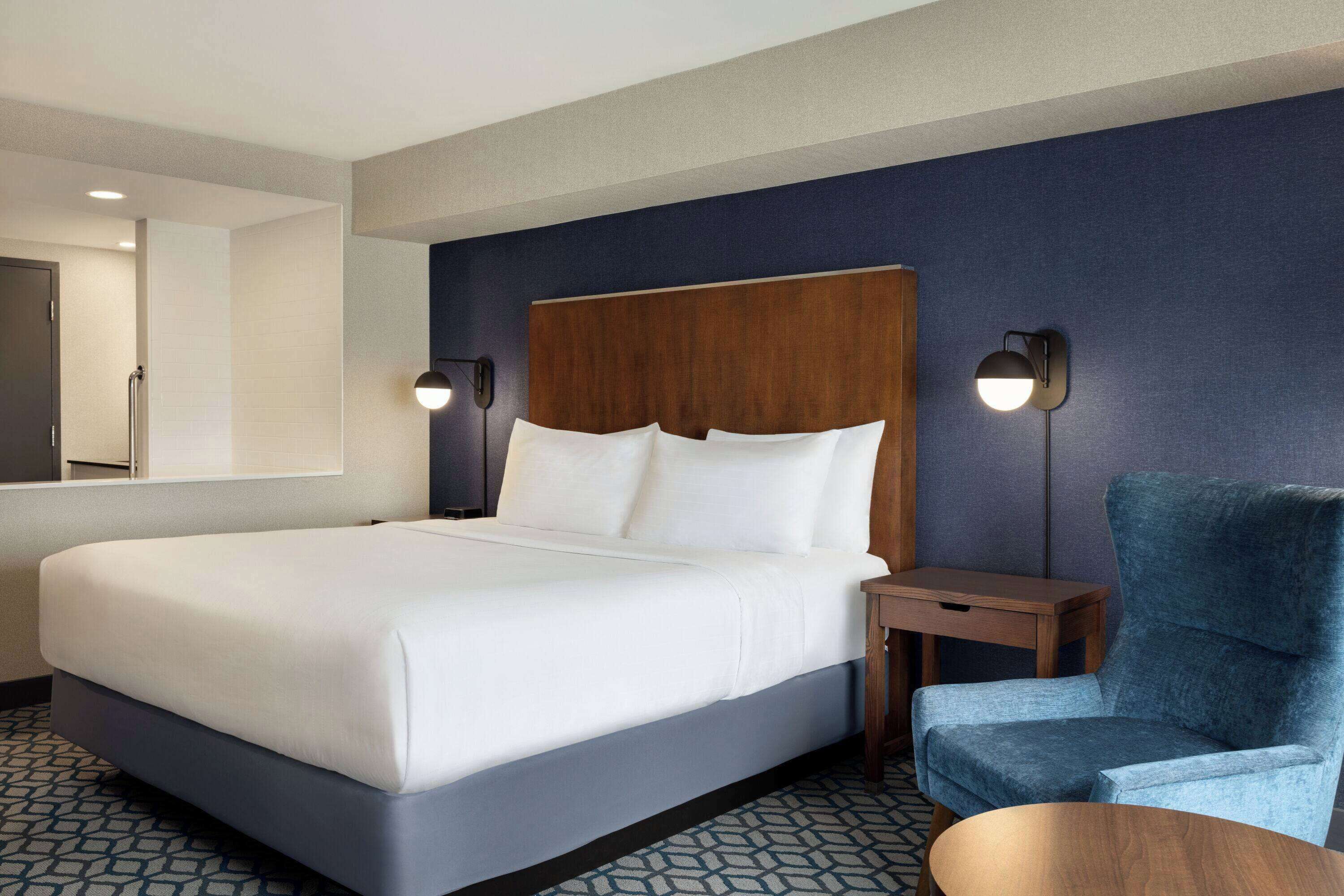 A hotel room with a large bed, a wooden headboard, two bedside lamps, a blue armchair, and a small wooden table.