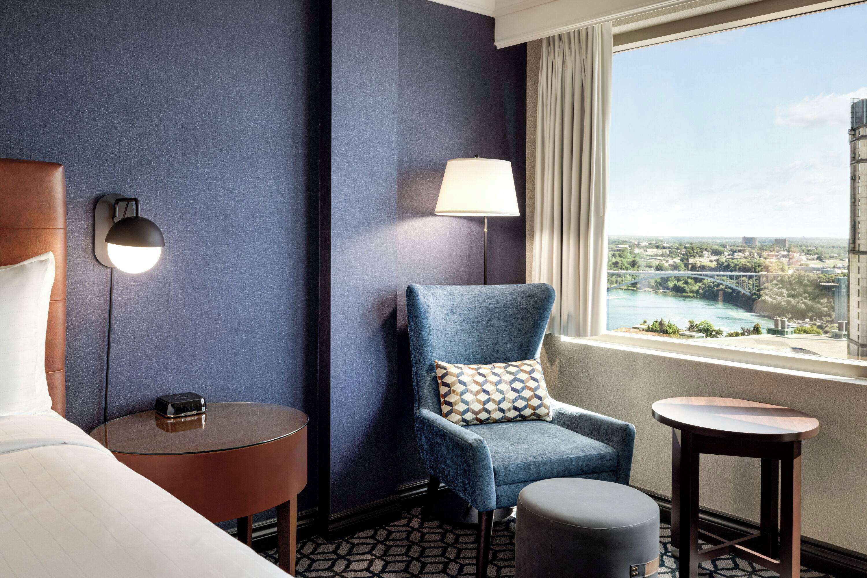 A hotel room with a bed, a blue armchair, a small round table, a lamp, and a view of a cityscape through a large window.