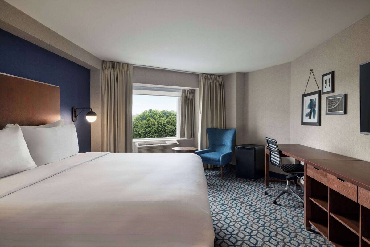 A hotel room with a large bed, a desk, a chair, and a view of greenery through the window.