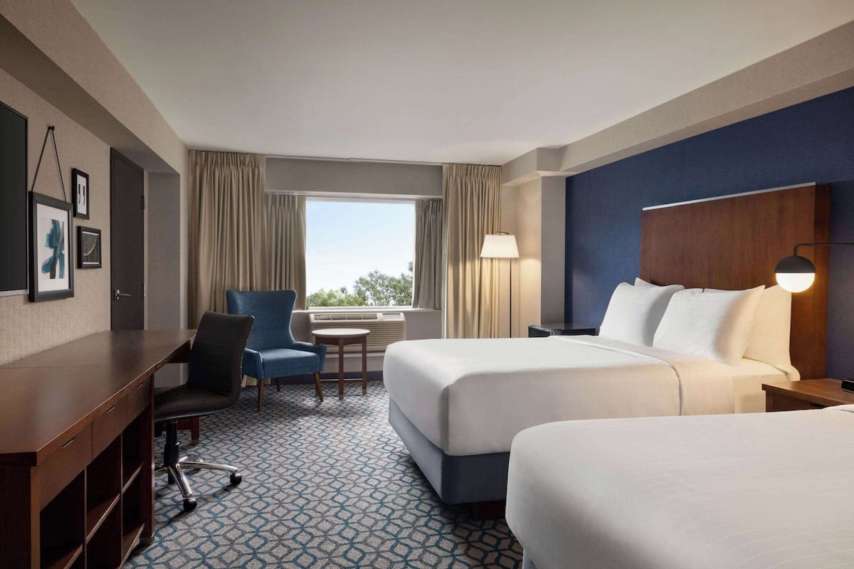 A hotel room with two beds, a desk, a chair, a lamp, and a window with curtains.