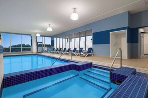 An indoor swimming pool with blue tiled edges, sunlit by natural light from large windows, and equipped with lounge chairs and a clear view of the outdoors.