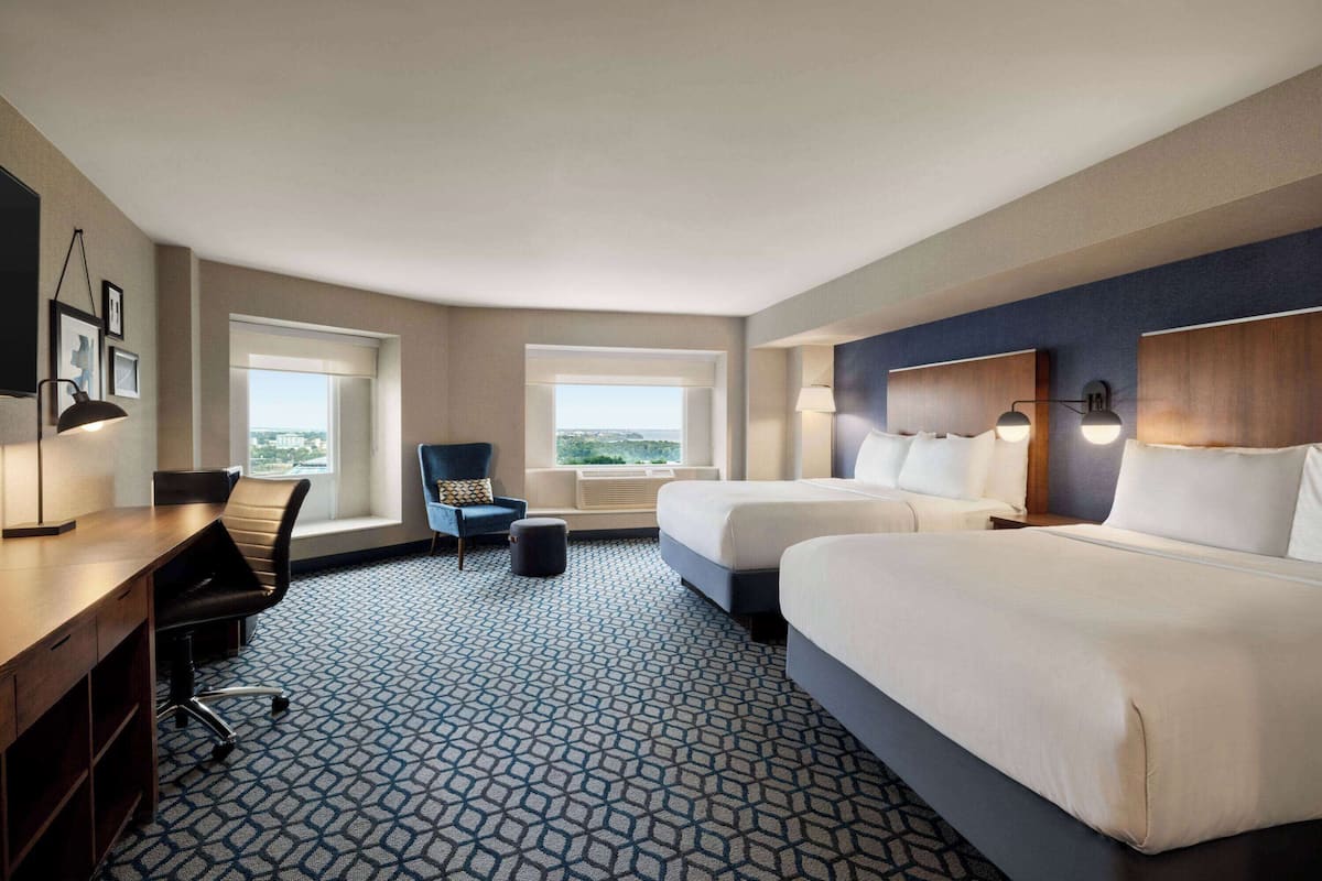 A hotel room with two beds, a desk, a chair, and large windows offering a city view.