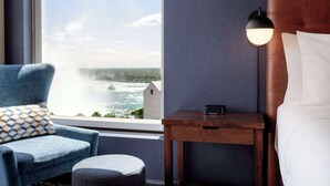 Une chambre d’hôtel avec vue sur un plan d’eau, un fauteuil bleu, une table de chevet en bois et un lit avec du linge de lit blanc.