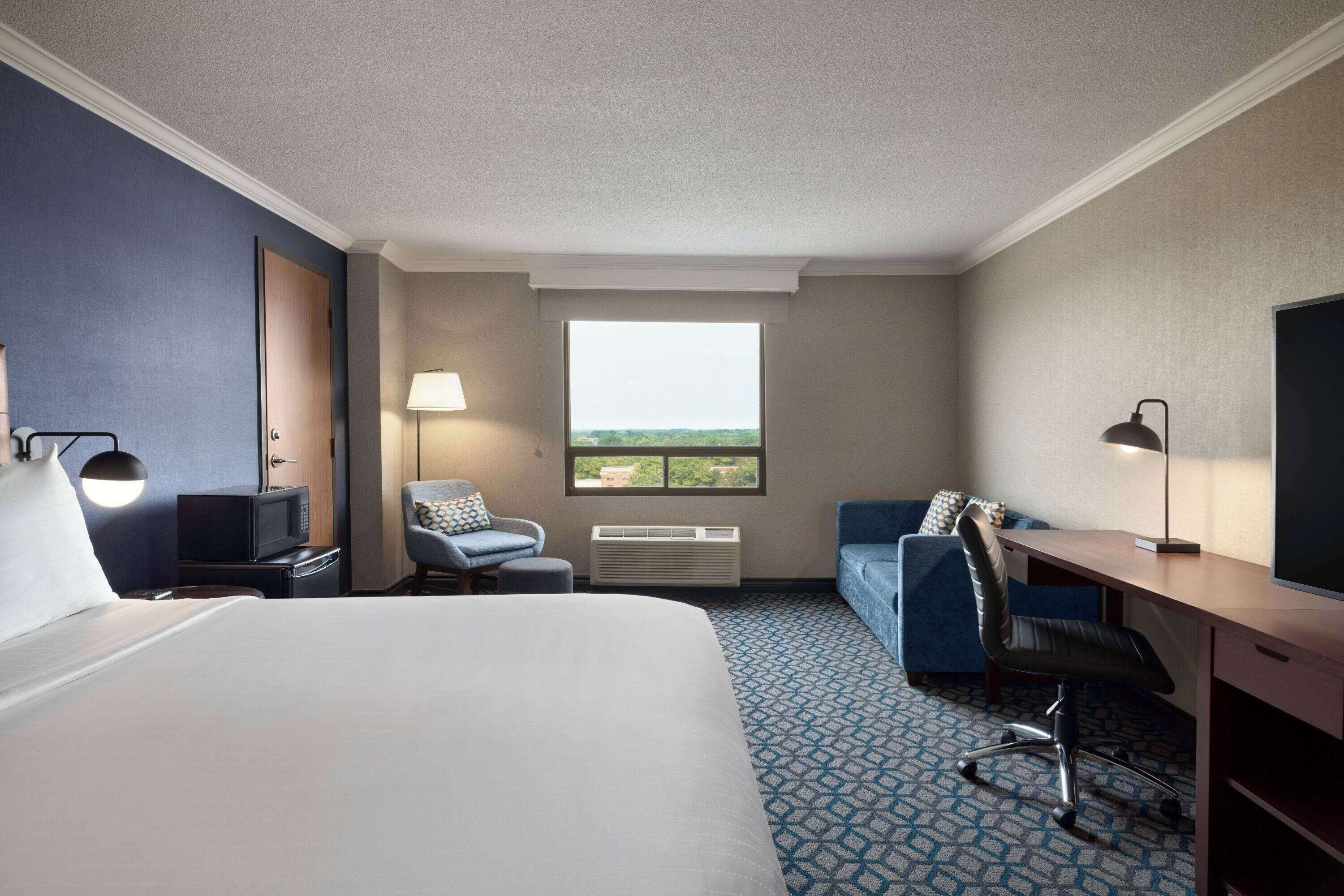 Une chambre d’hôtel avec un grand lit, un bureau, une chaise, une télévision, une lampe et une fenêtre donnant sur un beau paysage.