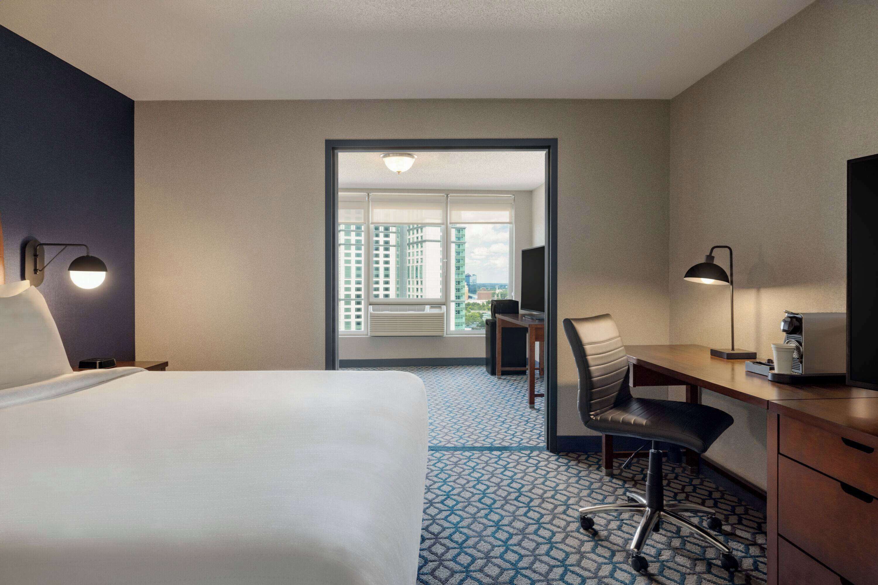 A hotel room with a bed, desk, chair, and a view of buildings through a door.