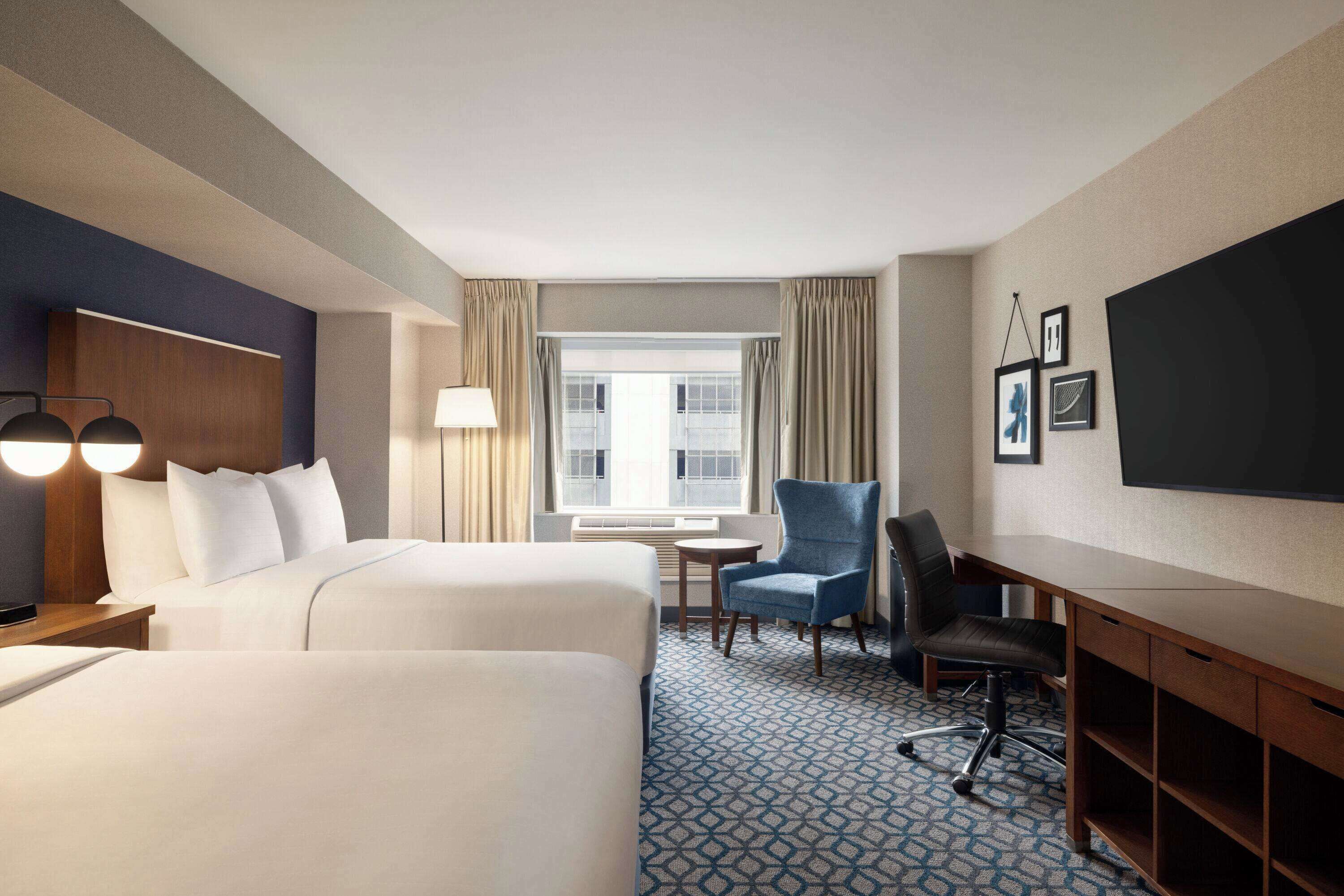 A hotel room with two beds, a desk, a chair, a TV, and a window with curtains.