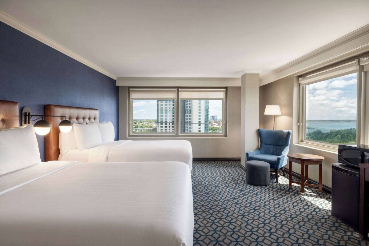 A hotel room with a large bed, a chair, a small table, and a TV. There are large windows with a view of buildings and a body of water.