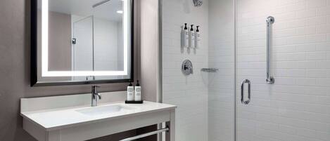 Un baño moderno con ducha envuelta en vidrio, un lavabo blanco con un espejo arriba y una repisa con toallas.