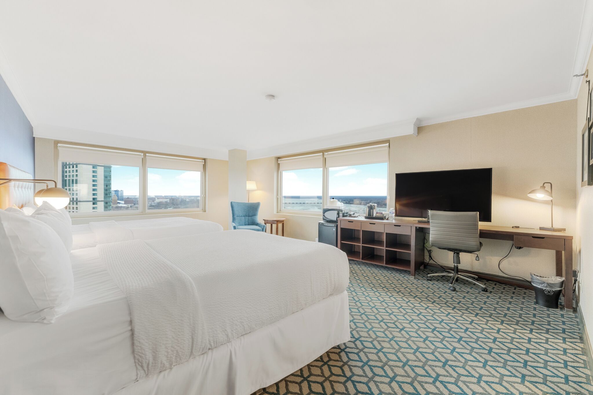 A hotel room with a large bed, a desk, a chair, a TV, and a view of the city.