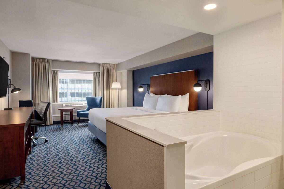 A hotel room with a large bed, a desk, a chair, a small table, a lamp, a window with curtains, a wall-mounted TV, and a bathtub.
