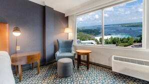 Une chambre d’hôtel dotée d’une grande fenêtre donnant sur une cascade et un pont. On y trouve un fauteuil bleu, une petite table ronde et un pouf gris.