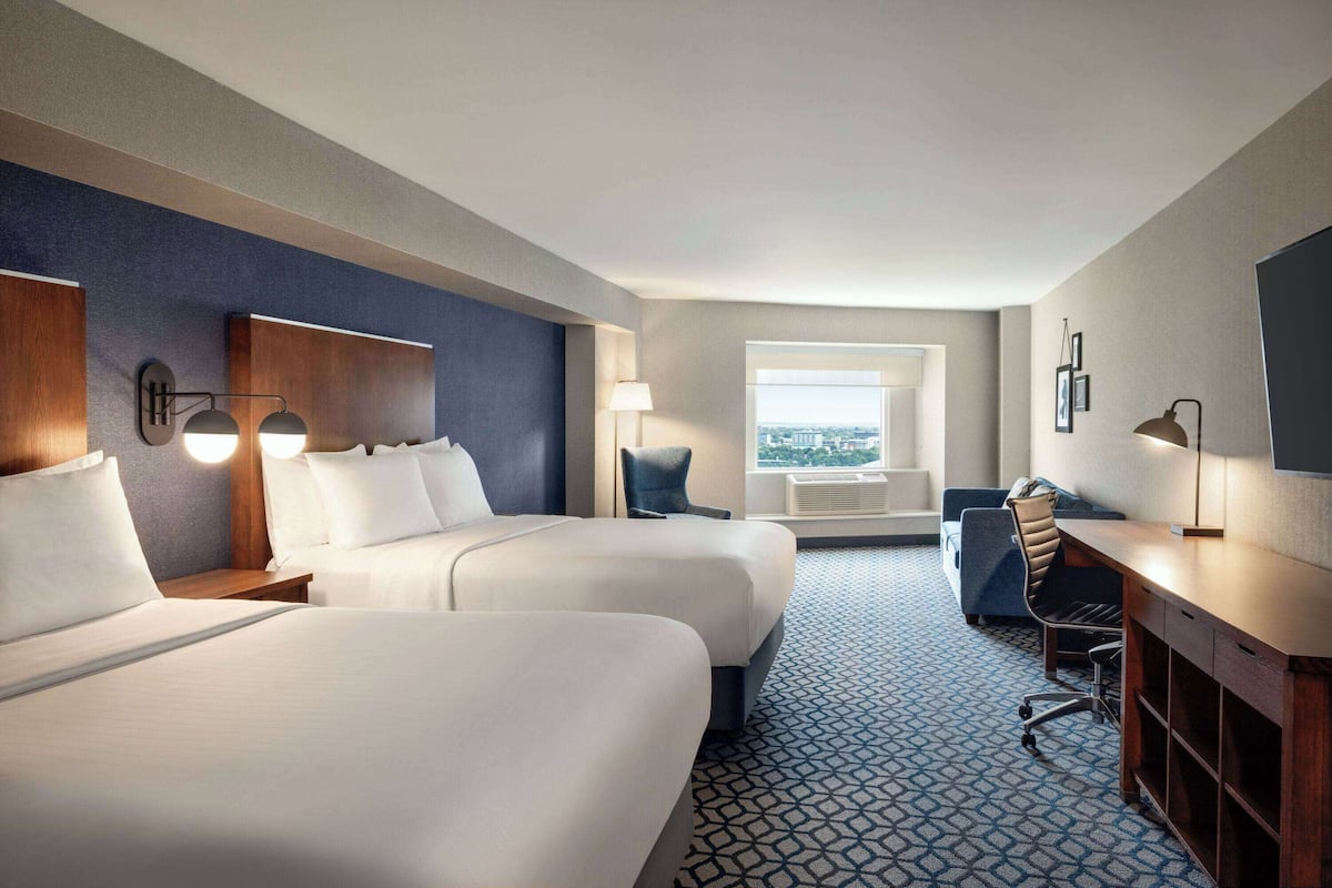 A hotel room with two beds, a desk, a chair, a lamp, and a window with a city view.