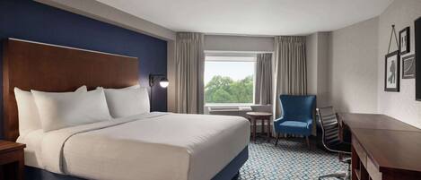 A hotel room with a large bed, a desk, a chair, and a view of trees through the window.