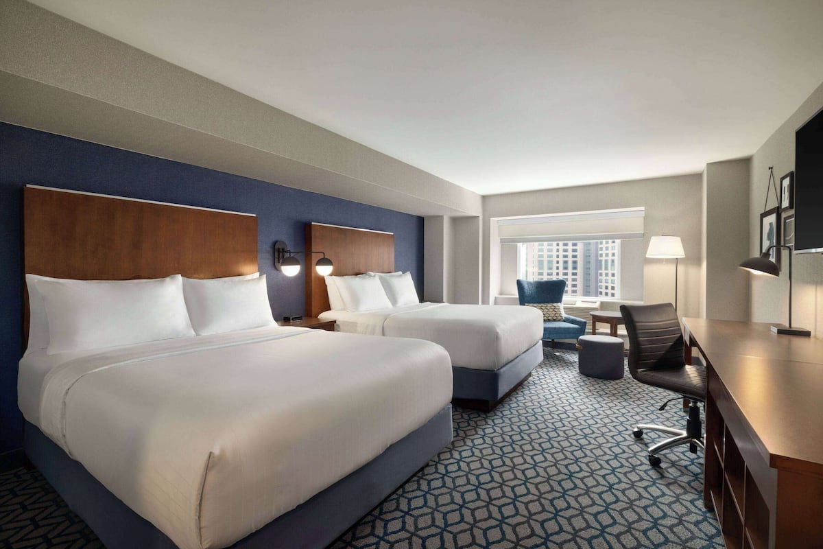A hotel room with two beds, a desk, a chair, and a window with a view of buildings.