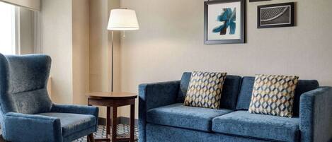 A room with a blue armchair, a sofa with patterned cushions, a small round table, and a floor lamp. There are framed artworks on the wall.
