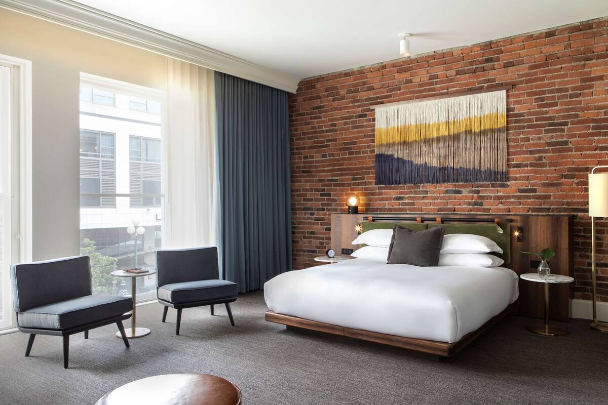 Une chambre d’hôtel avec un mur en briques, un grand lit, deux fauteuils, une petite table et une vue sur les bâtiments environnants.