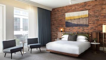 Habitación de hotel con pared de ladrillo, una cama grande, dos sillones, una mesita y vistas a edificios.