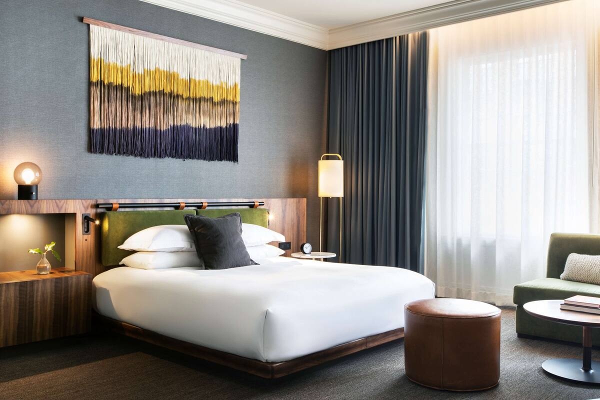 Une chambre d’hôtel avec un grand lit, une table de chevet avec une lampe, un fauteuil vert recouvert de tissu, un pouf rond et une œuvre d’art abstraite accrochée au mur.
