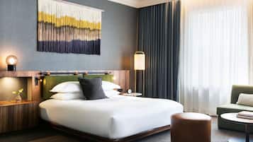 A hotel room with a large bed, a bedside table with a lamp, a green upholstered chair, a round ottoman, and a wall-mounted abstract art piece.