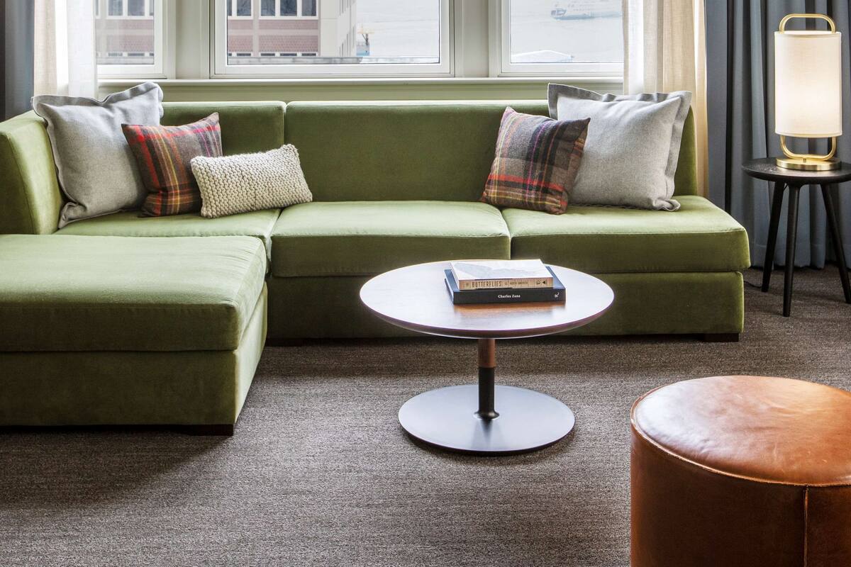 Un canapé vert agrémenté de plusieurs coussins, une table basse ronde sur laquelle repose un livre, et une vue sur un bâtiment visible à travers la fenêtre.