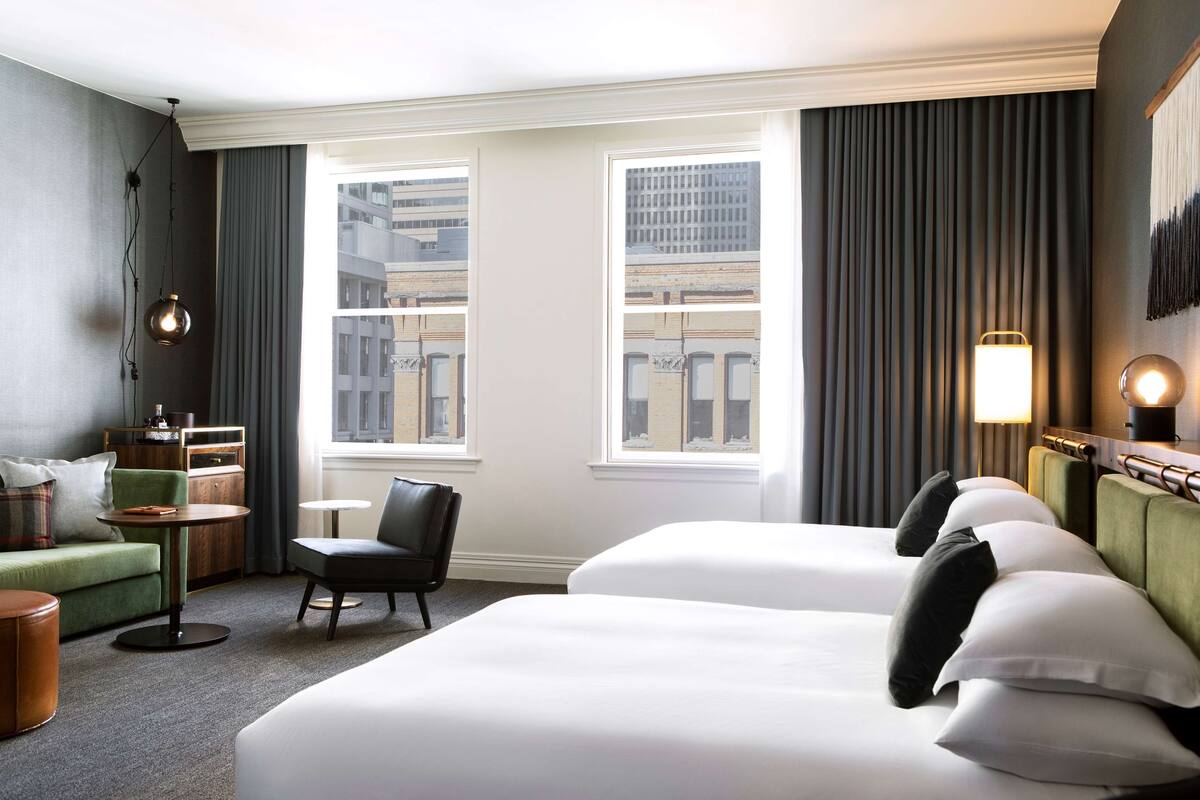 Une chambre d’hôtel avec deux lits, un canapé vert, une petite table et une chaise. Il y a des fenêtres avec des rideaux, une lampe sur la table de chevet et une vue sur les bâtiments à l’extérieur.