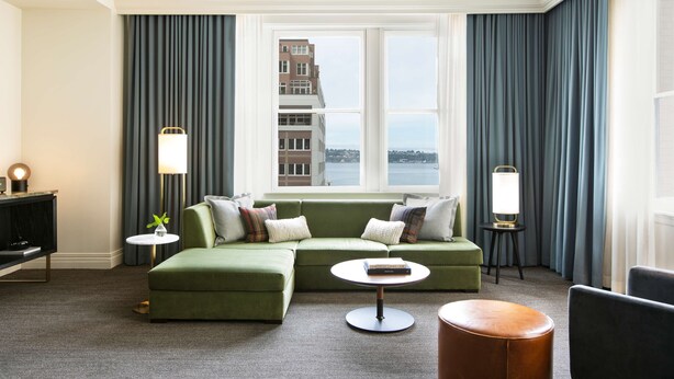 Une chambre d’hôtel moderne avec un canapé d’angle vert, une table basse ronde, un pouf en cuir et une vue sur le paysage urbain à travers de grandes fenêtres.