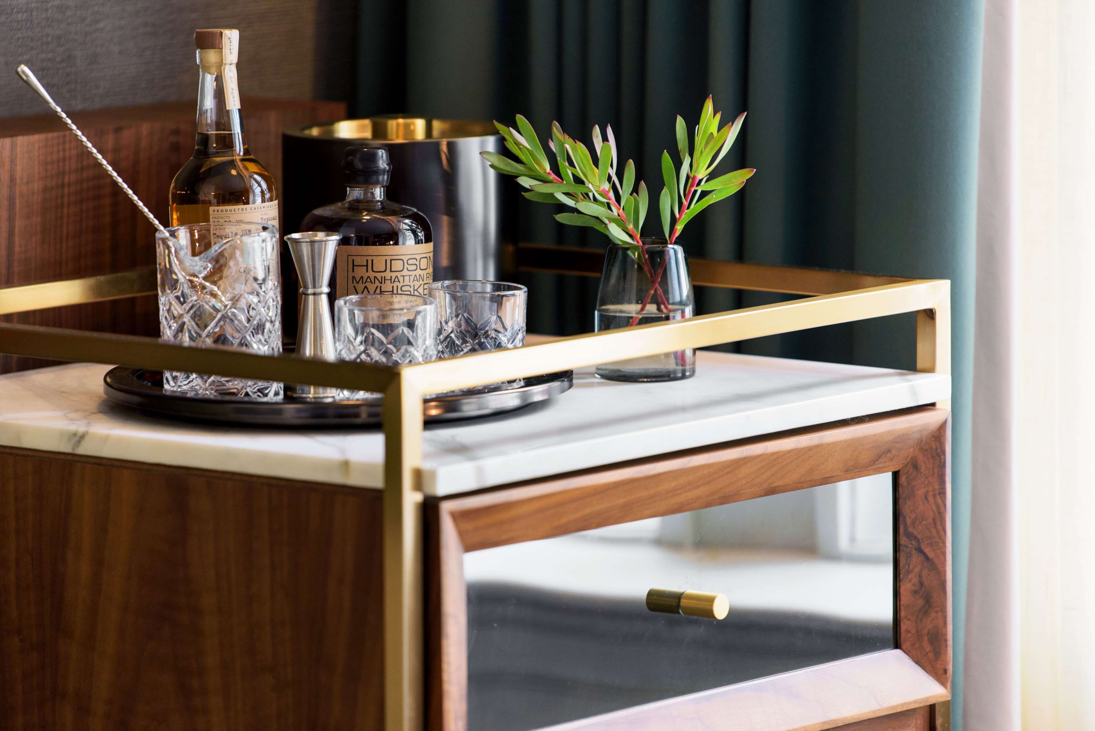 A bar cart with a bottle of Hudson Manhattan, a glass, and a small vase with green leaves.