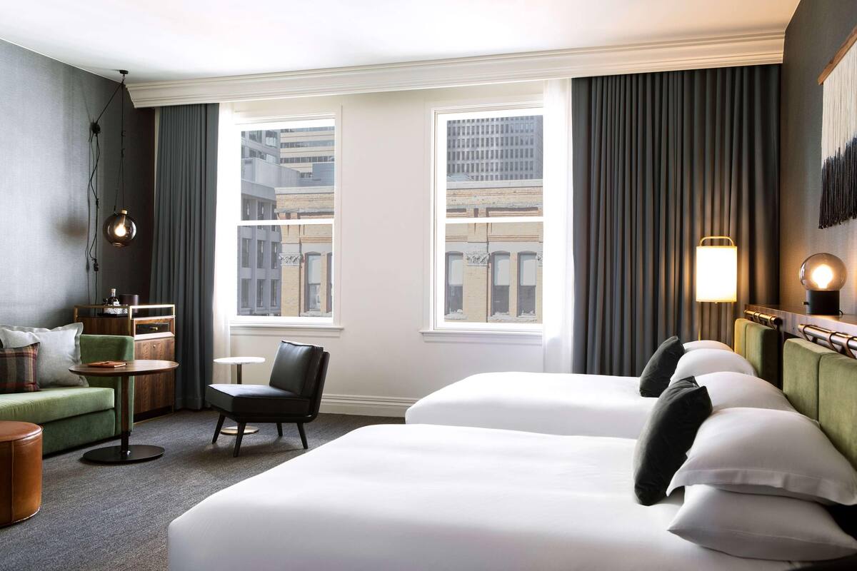 Une chambre d’hôtel avec deux lits, un canapé vert, une chaise, une petite table et une vue sur les immeubles à travers de grandes fenêtres.
