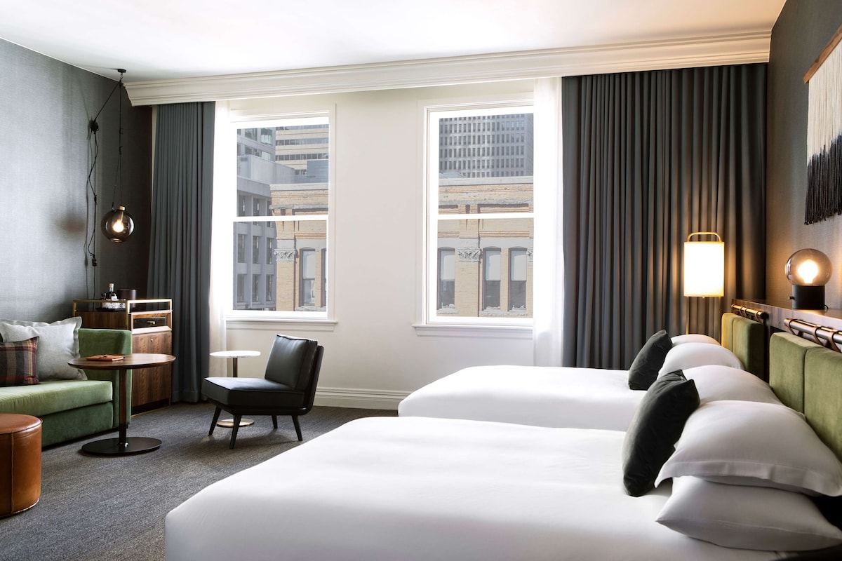 Une chambre d’hôtel avec deux lits, un canapé vert, une petite table et une chaise. Il y a des fenêtres avec des rideaux, une lampe sur la table de chevet et une vue sur les bâtiments à l’extérieur.