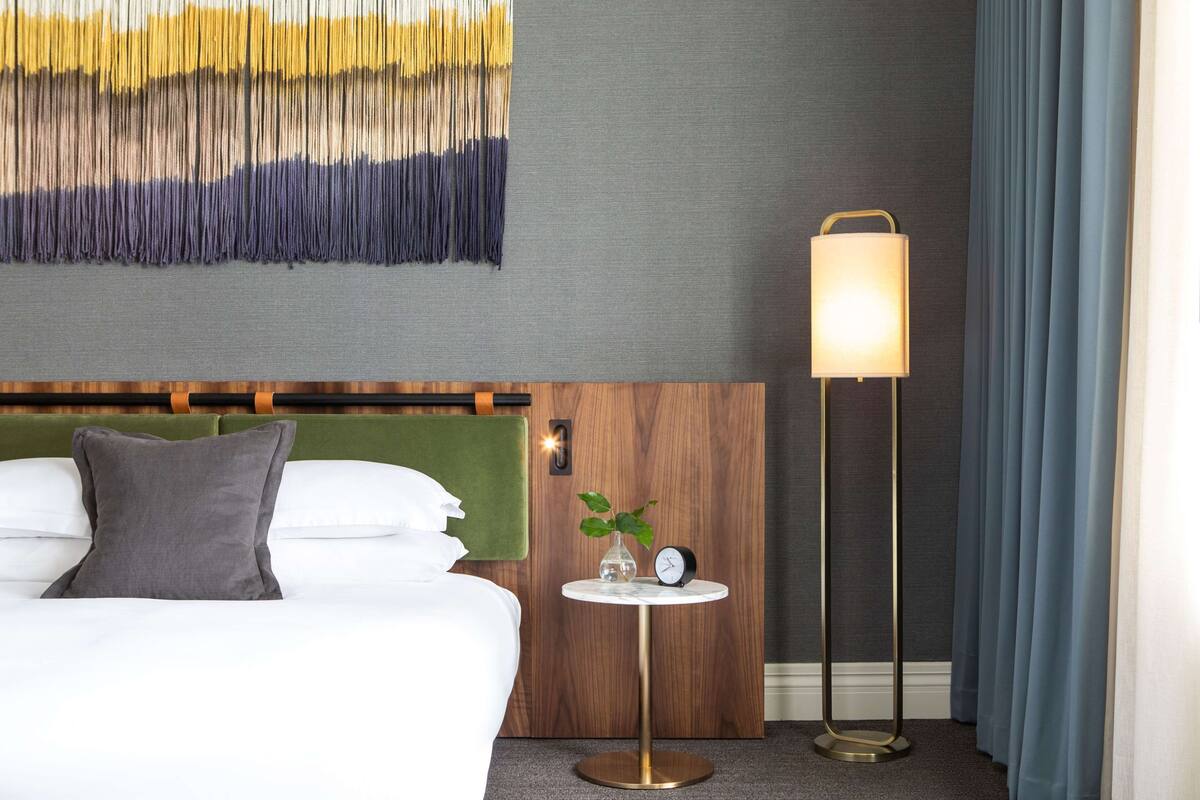 Une chambre d’hôtel avec un lit, une petite table ronde avec une horloge et une plante, une lampe fixée au mur et une tête de lit ornée d’un panneau en tissu vert.