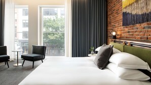 Une chambre d’hôtel avec un grand lit, une tête de lit verte, une petite table avec une lampe, une chaise et une vue sur la ville grâce à de grandes fenêtres.