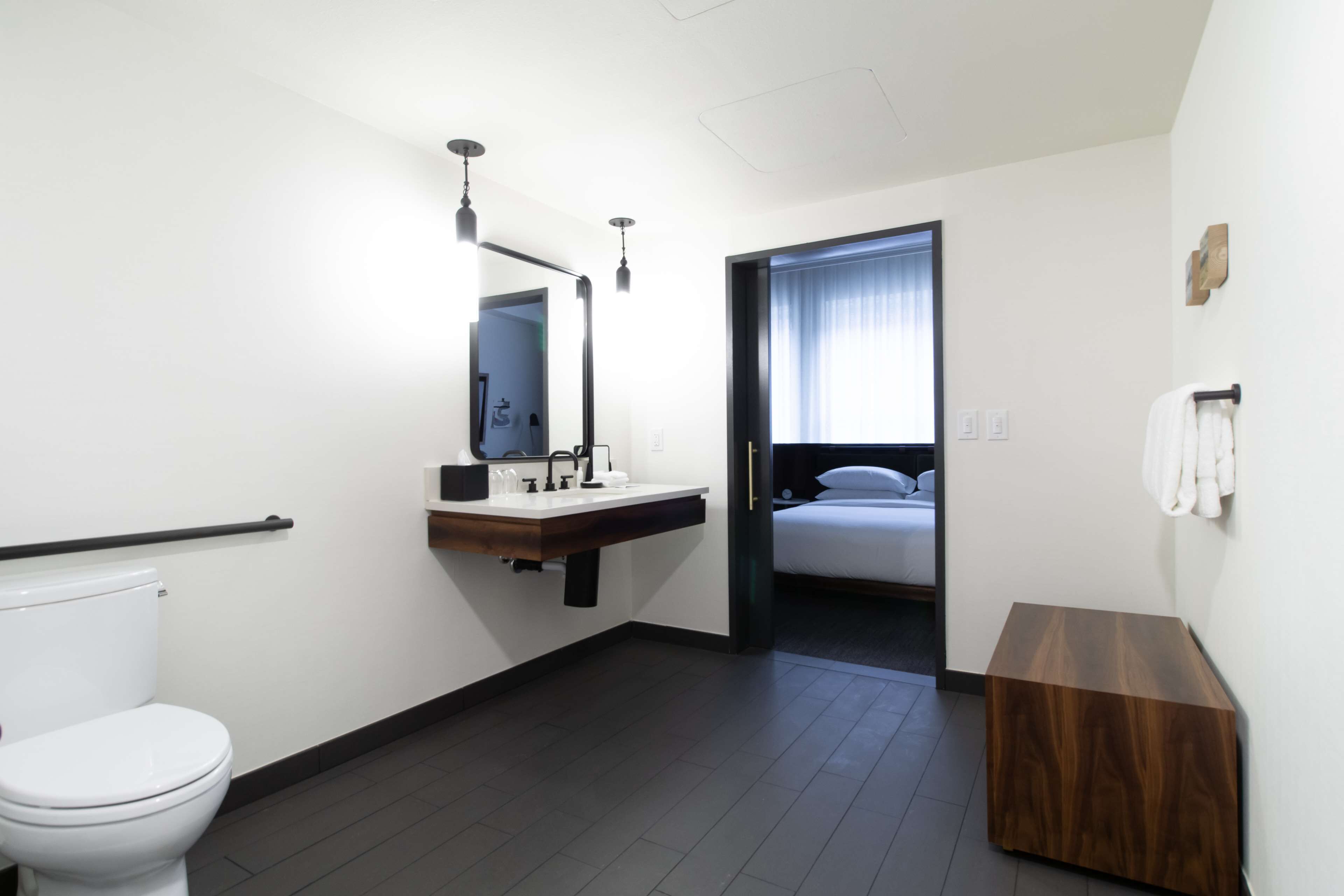 A modern bathroom with a toilet, sink, and mirror. There is a wooden bench and a towel rack on the wall.