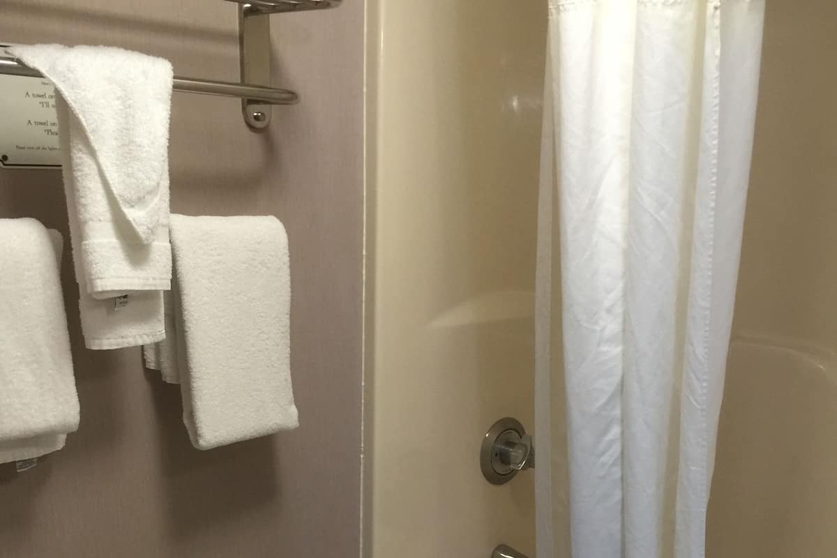 Uma casa de banho com sanita, um rolo de papel higiénico verde, uma banheira com cortina de chuveiro branca e várias toalhas brancas penduradas num suporte.