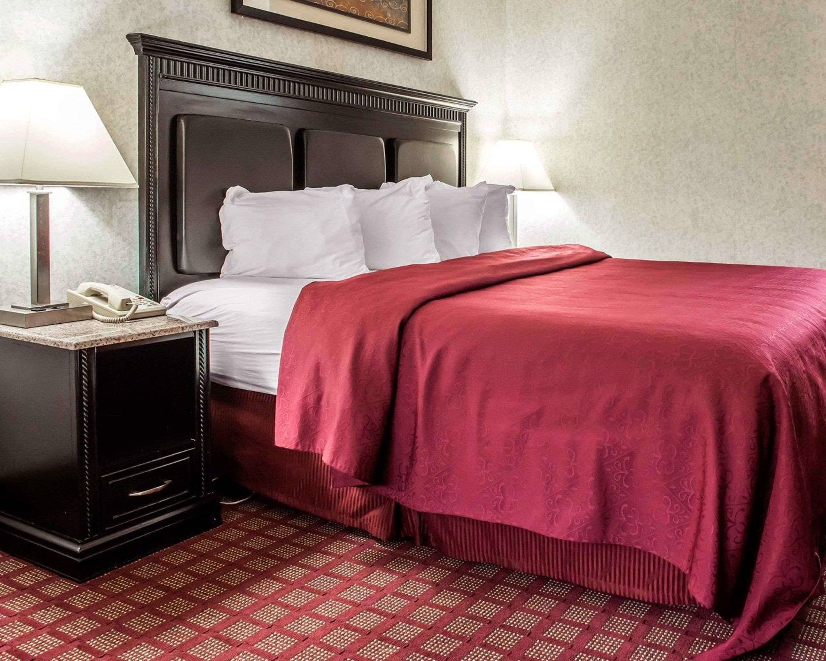 A hotel room with a dark wood headboard, white bedding, a red throw blanket, a telephone on a nightstand, and a patterned red carpet.
