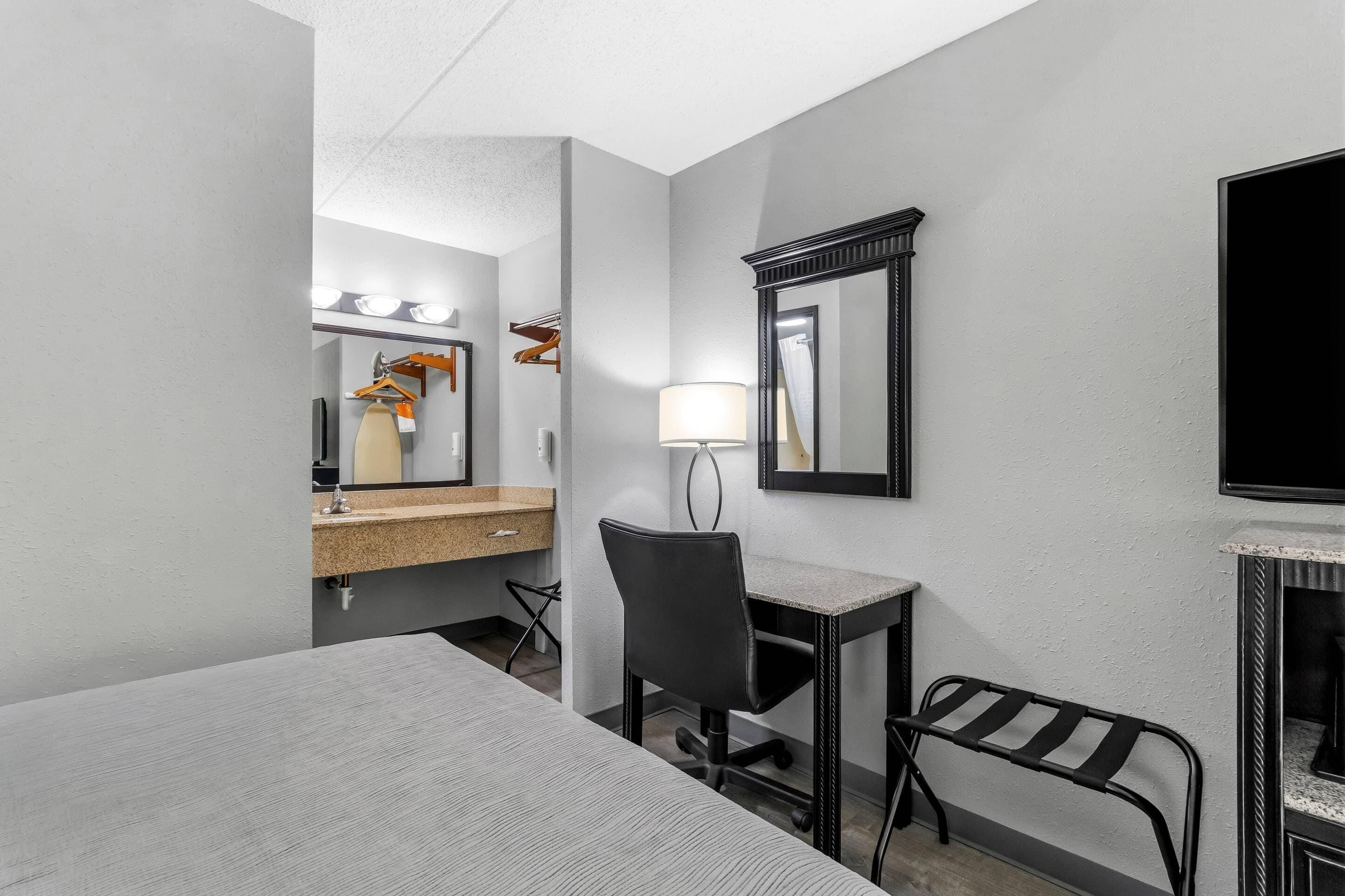 A hotel room with a bed, a desk, a chair, a mirror, a lamp, and a television.