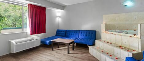 Une pièce moderne comprenant un canapé d’angle, une petite table, un comptoir de cuisine intégré et une fenêtre avec des rideaux rouges.