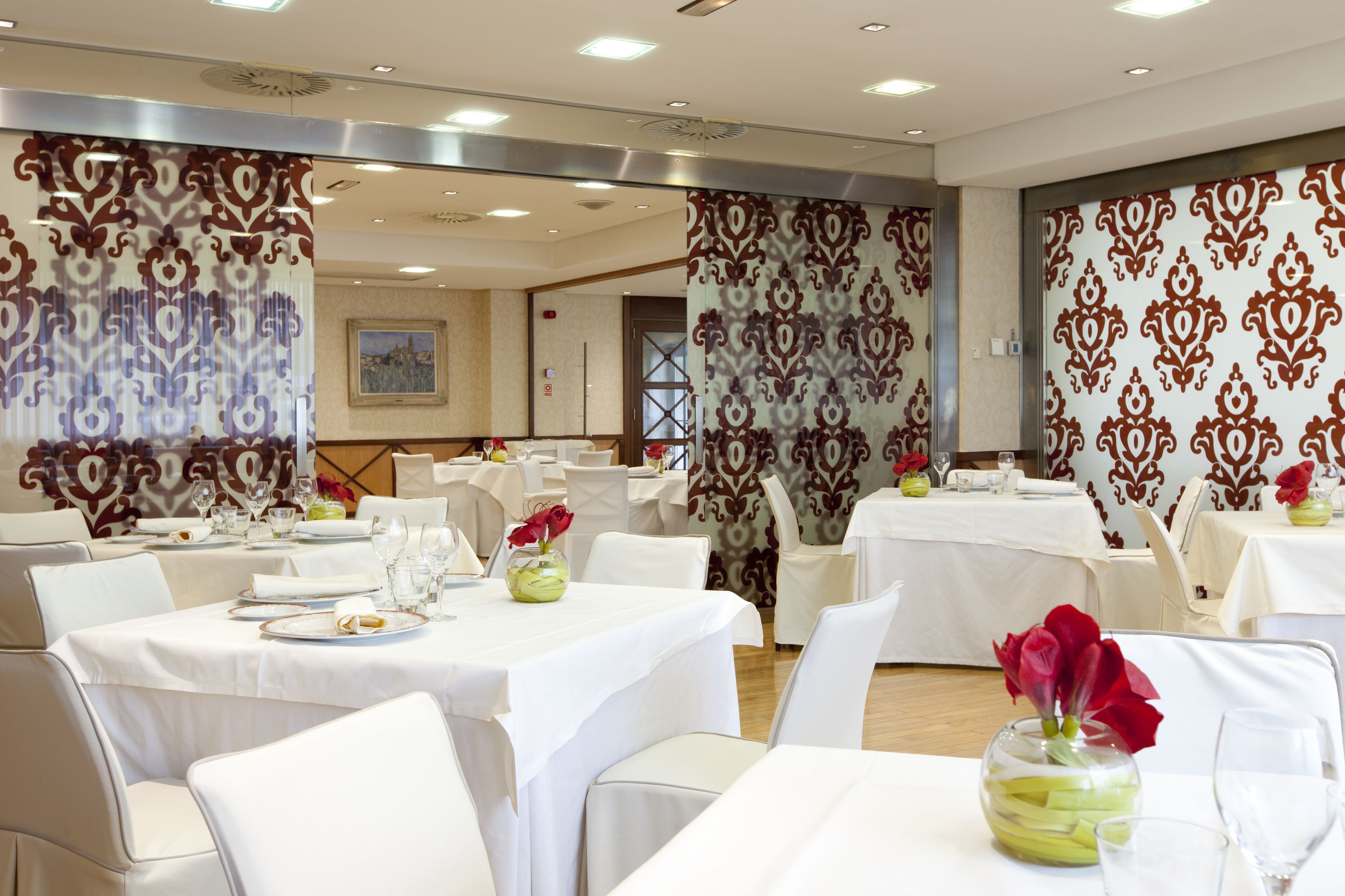 Comedor de hotel con varias mesas dispuestas para los huéspedes, con manteles blancos y flores rojas en jarrones.