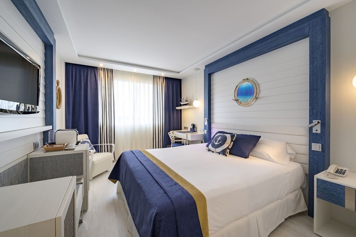 A hotel room with a bed, bedside table, desk, chair, and a large window with curtains.