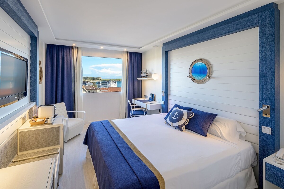 A hotel room with a bed, a desk, a chair, a TV, and a view of the city.